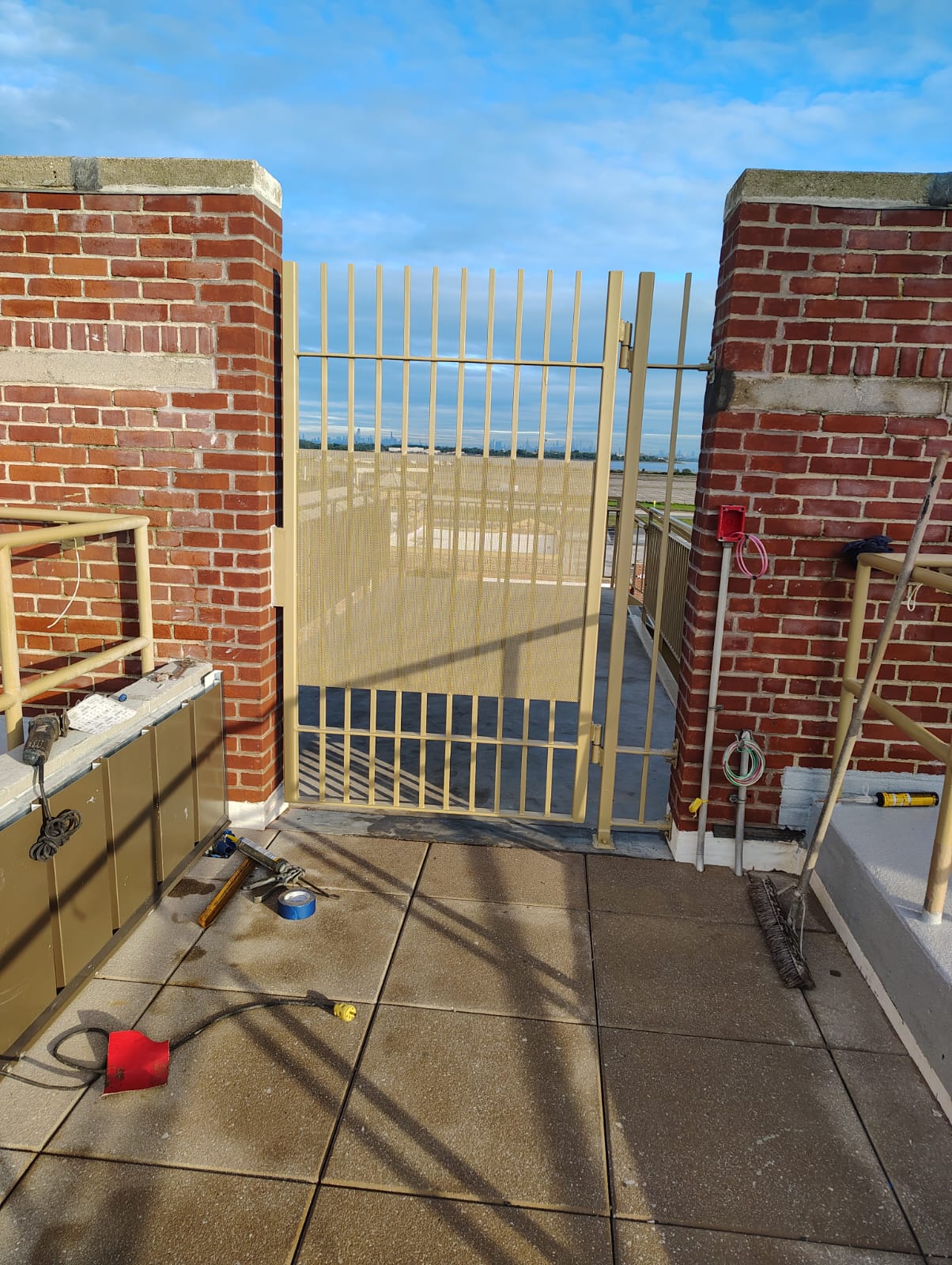 Painted metal access gate with vertical bars and mesh infill between brick piers